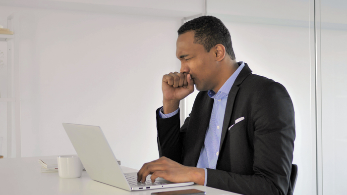 Man in a suit sitting at a desk with a laptop, coughing into his hand