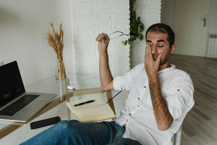 Man sitting at a desk with a laptop, holding glasses and looking tired.
